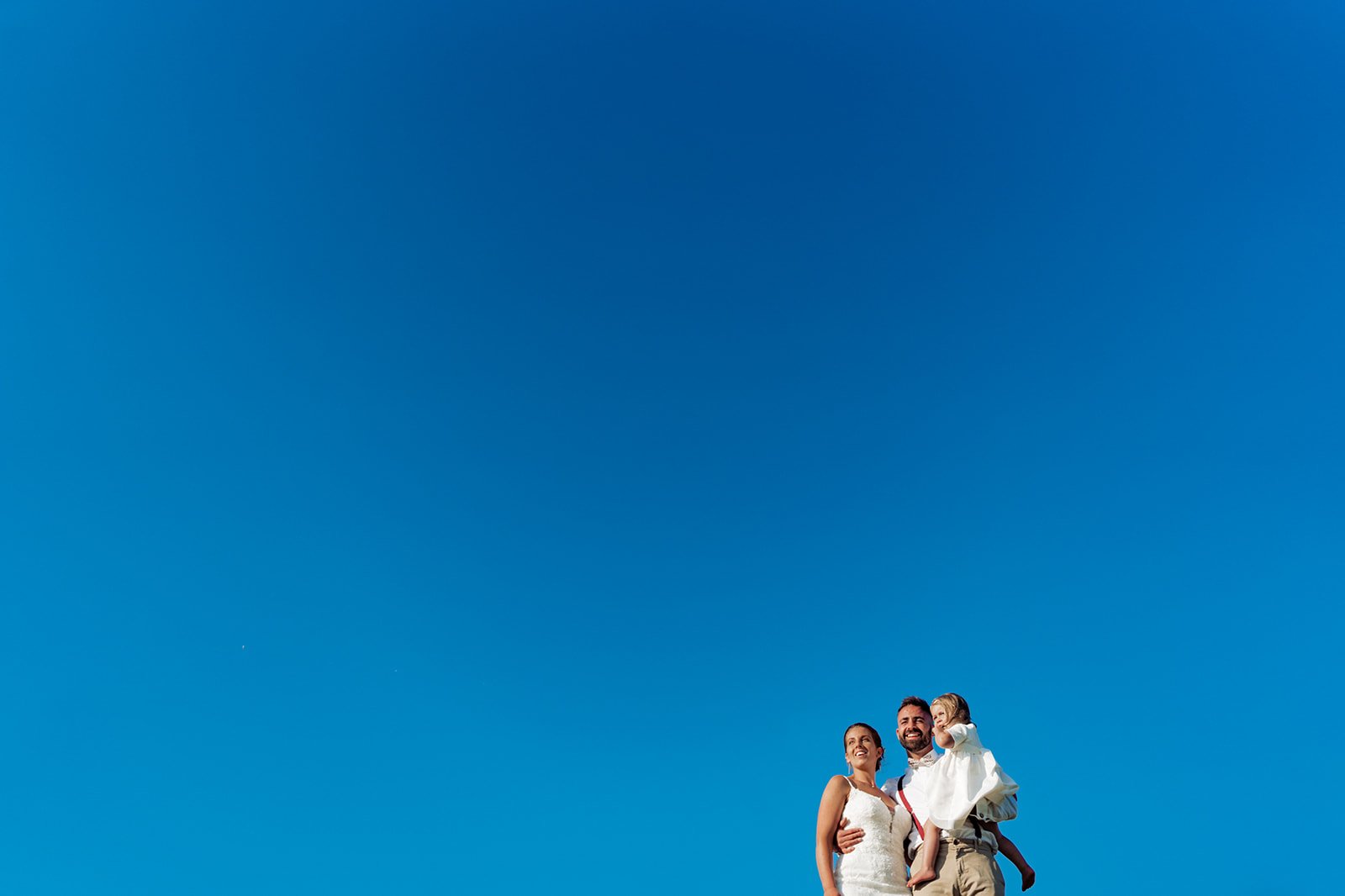 Happy wedding family portrait against a bright blue sky. Bride, groom, and child. North Devon wedding photography.