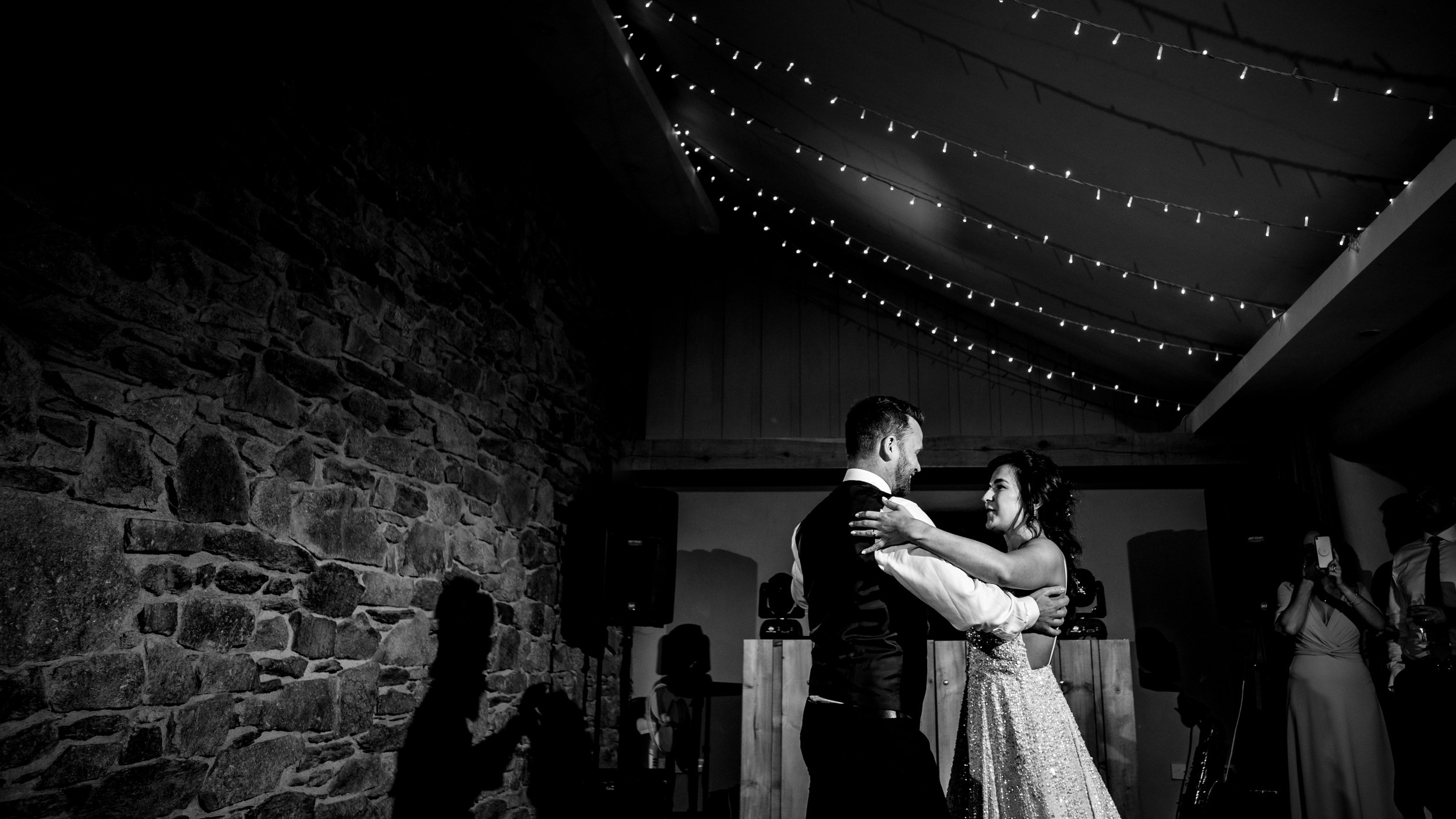 Black and white wedding photo of bride and groom dancing under string lights. Stone wall in background.