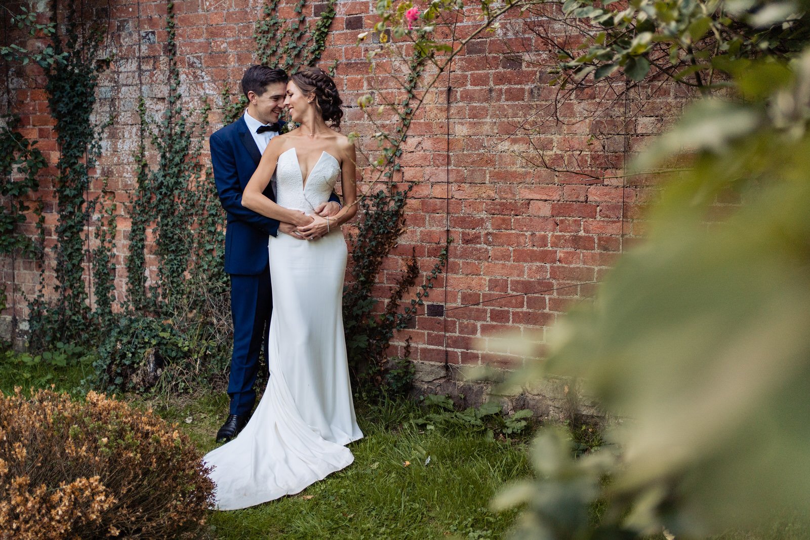 Bride and groom embrace in garden setting. Wedding portrait with brick wall backdrop. Elegant wedding attire. Bride and groom embrace in garden setting. Wedding portrait with brick wall backdrop. Elegant wedding attire.