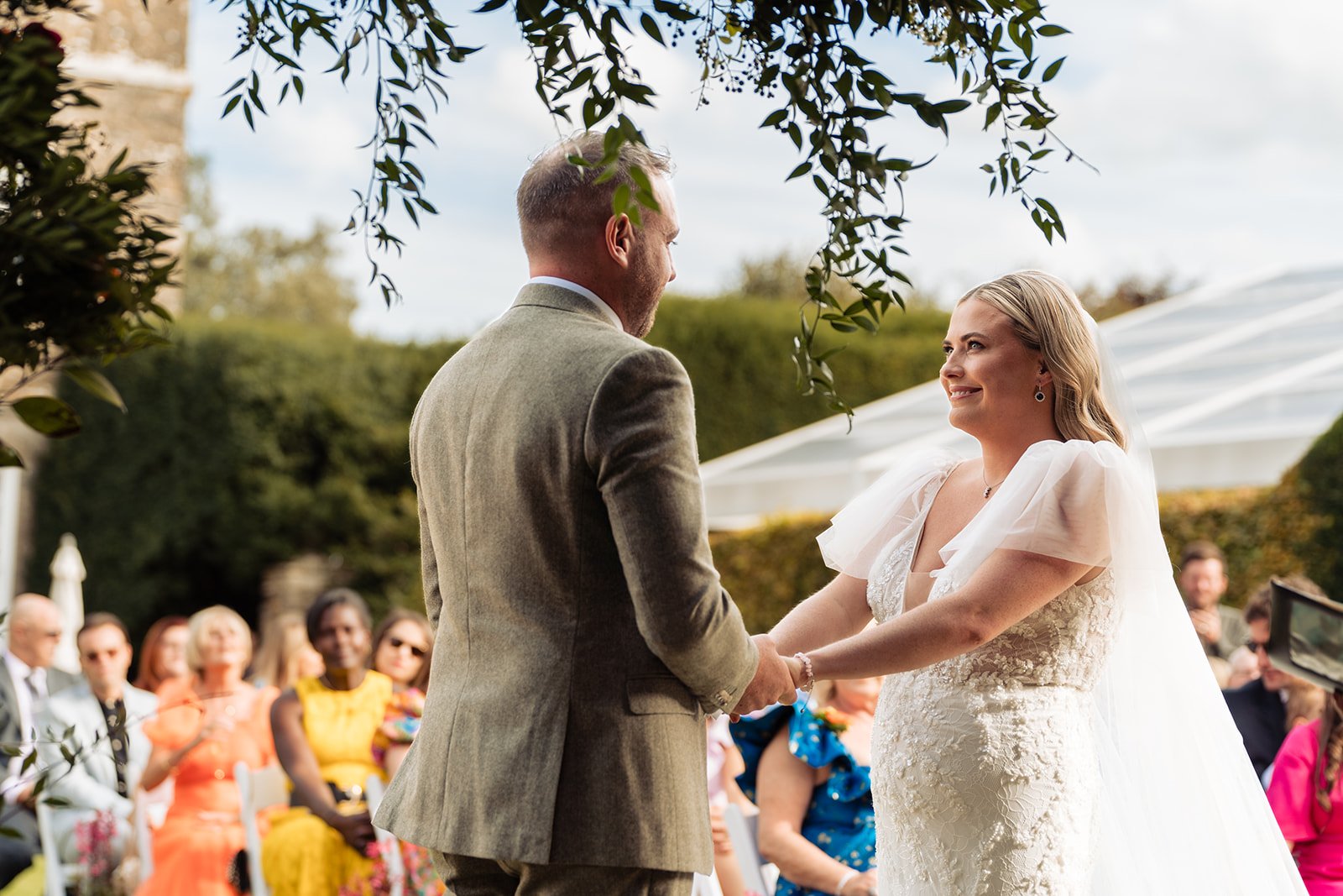 Bride and groom holding hands at outdoor wedding ceremony. Wedding guests in background.