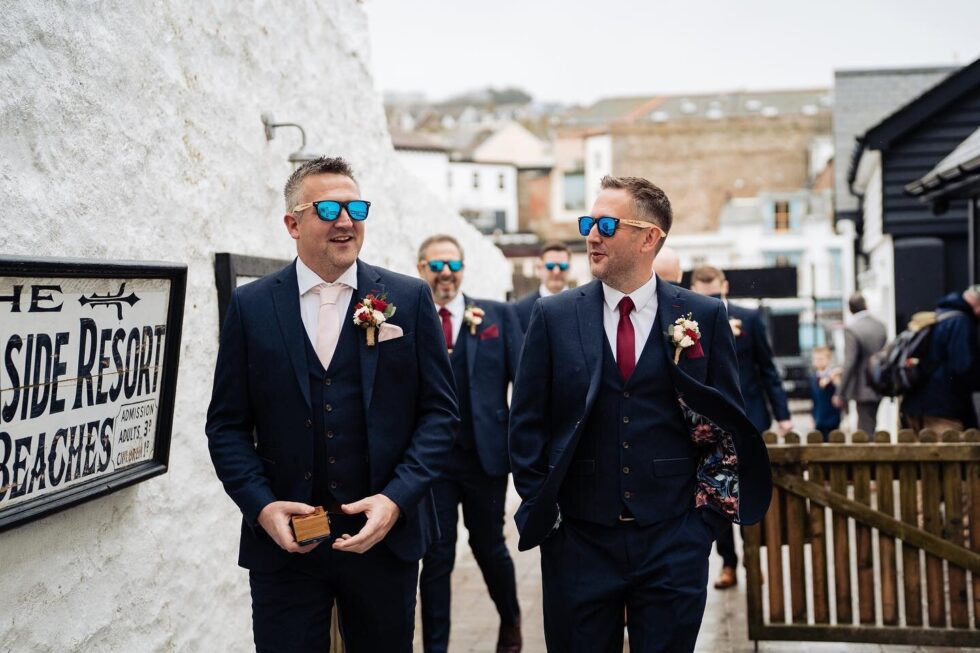 Groomsmen in suits and sunglasses walk to a wedding. Seaside resort in background. Beach wedding party.