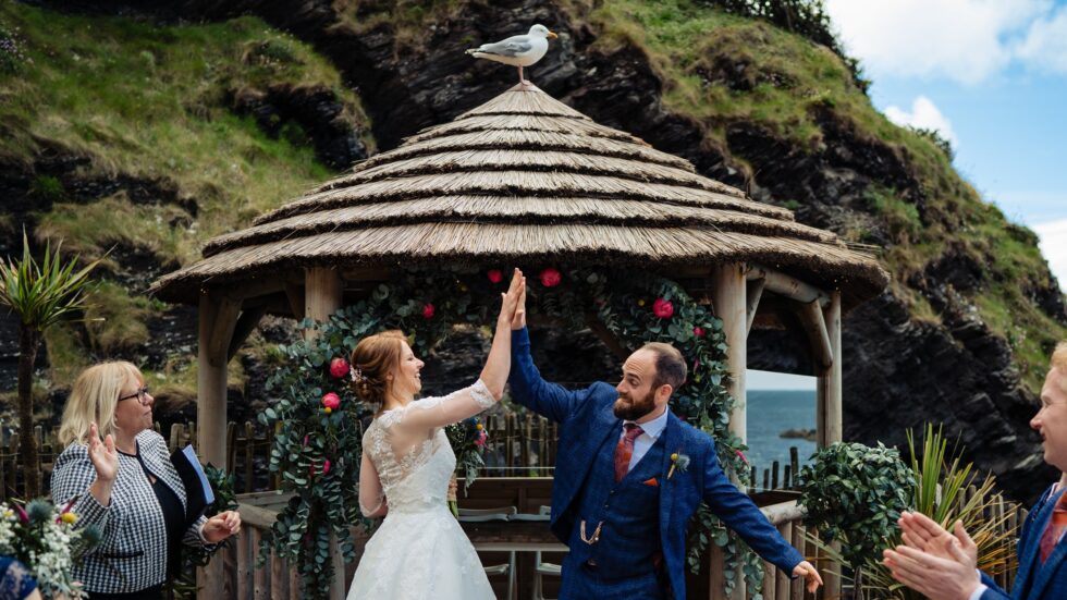 Devon wedding photography: Bride and groom high-five under gazebo with seagull on top. Tunnels Beaches wedding.