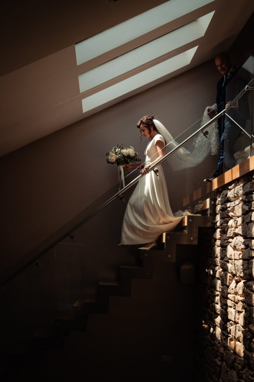 Cornwall wedding photo: Bride with flowers descends stairs at Trevenna Barns, sunlight above. Yeti Wedding Photography.
