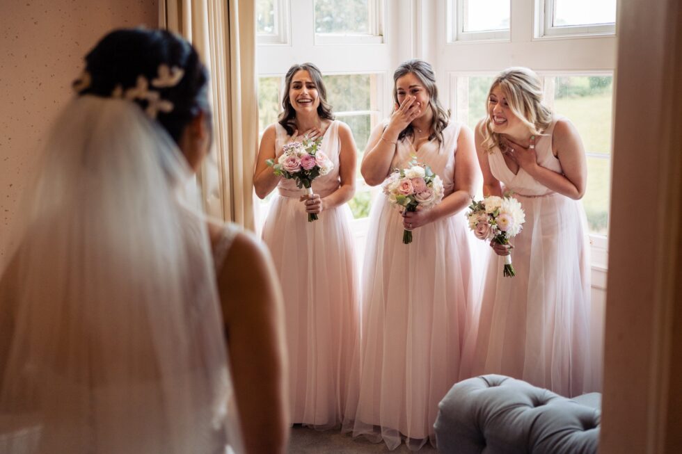 Bride in veil, bridesmaids in pink dresses react with bouquets. Devon wedding photography.