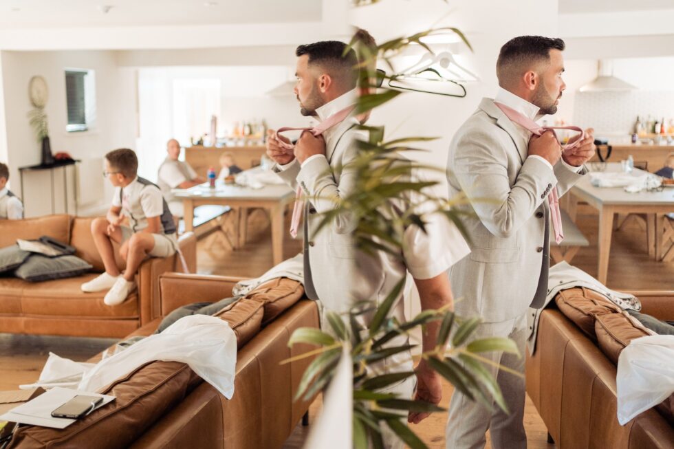 Groom getting ready for his Devon wedding. Groom in suit tying tie, wedding party in background. Yeti Wedding Photography.