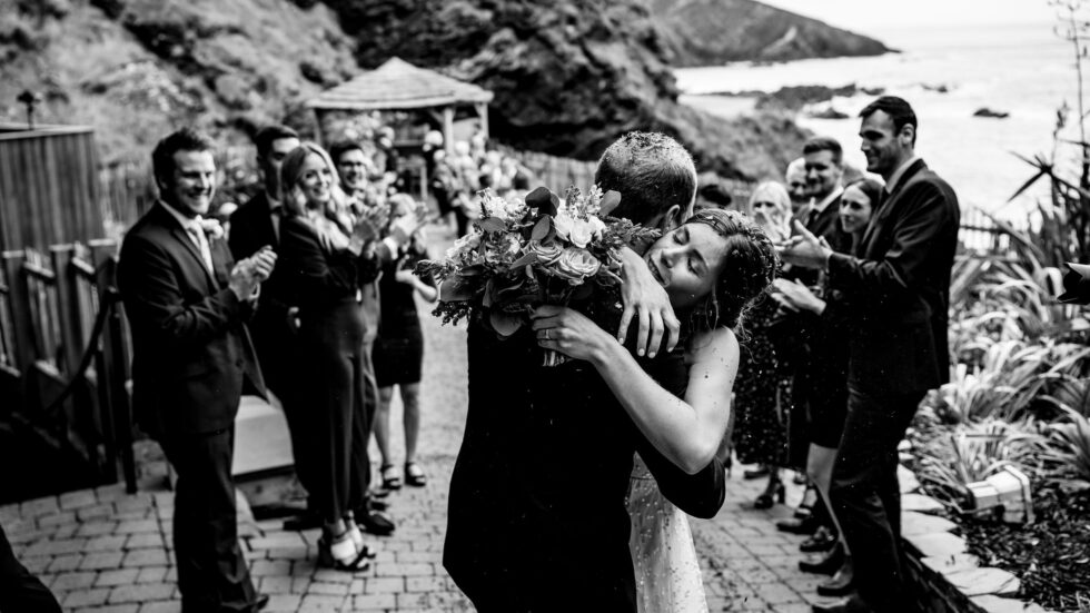 Bride and groom hug at Tunnels Beaches wedding in Devon. Guests applaud. Yeti Wedding Photography captures the joy.