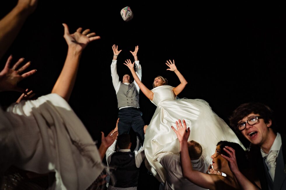 Rugby wedding photo: Bride and groom lifted by guests reaching for rugby ball. Yeti Wedding Photography. Huntsham Court wedding.