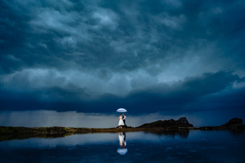 Stormy wedding photo: Bride and groom under umbrella, reflected in water. Dramatic sky.