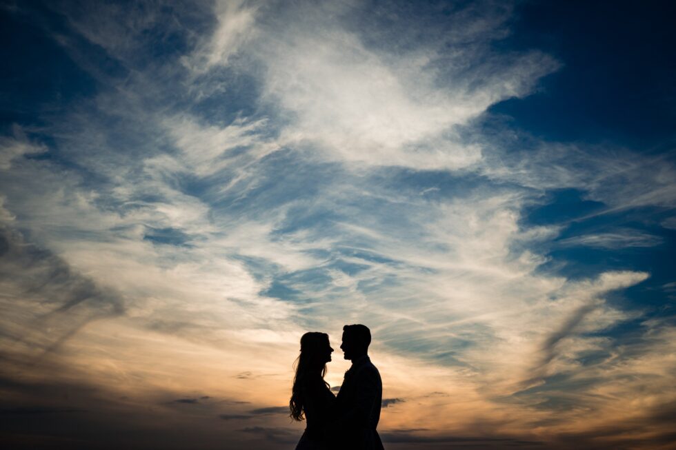 Devon wedding photography: Silhouette of bride and groom against a dramatic sunset sky with clouds.