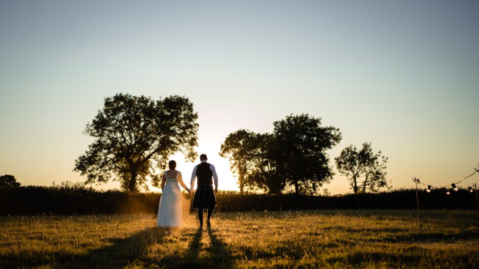 Sunset wedding photo of a bride and groom holding hands in a field. Devon wedding photography.