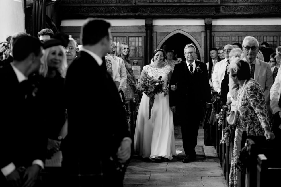 Devon wedding photography: Bride walks down aisle with father. Black and white image of wedding ceremony.