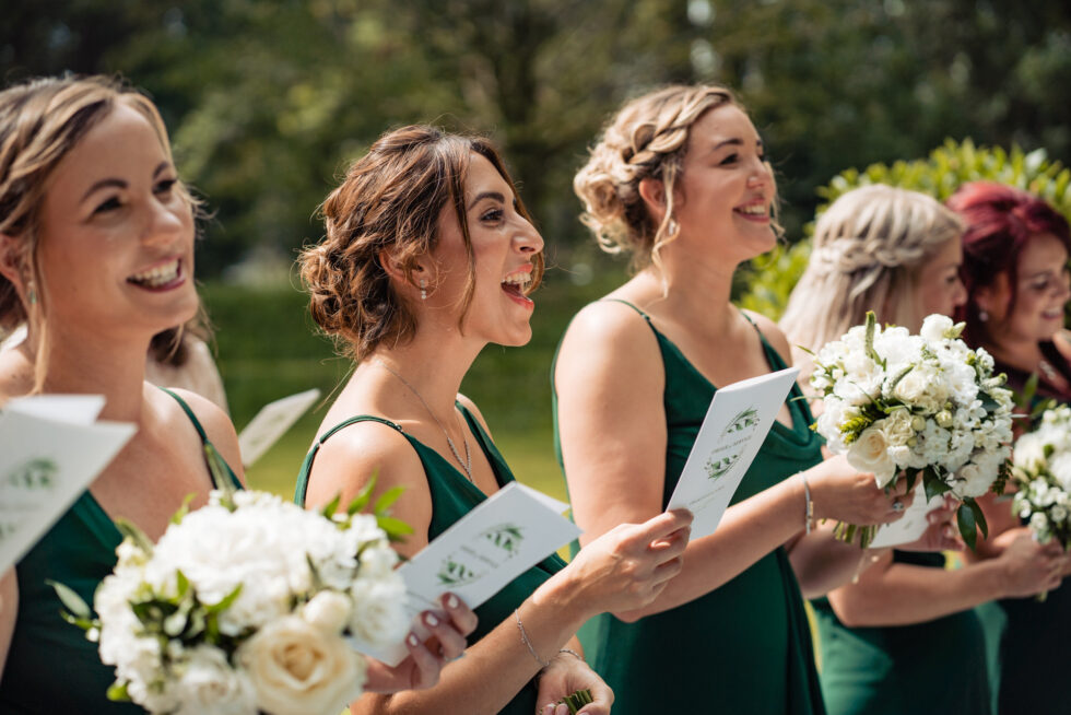Bridesmaids in green dresses sing at a wedding ceremony. They hold bouquets and song sheets.