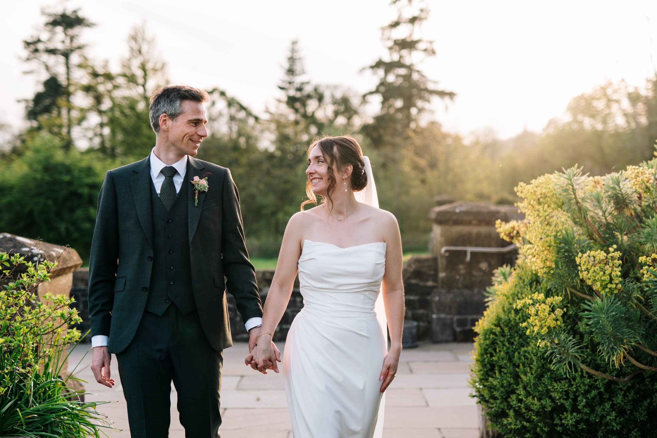 Walking hand in hand, Elsa & Paul at Huntsham Court Wedding in Devon, lit by warm golden-hour light with greenery and elegant suits.