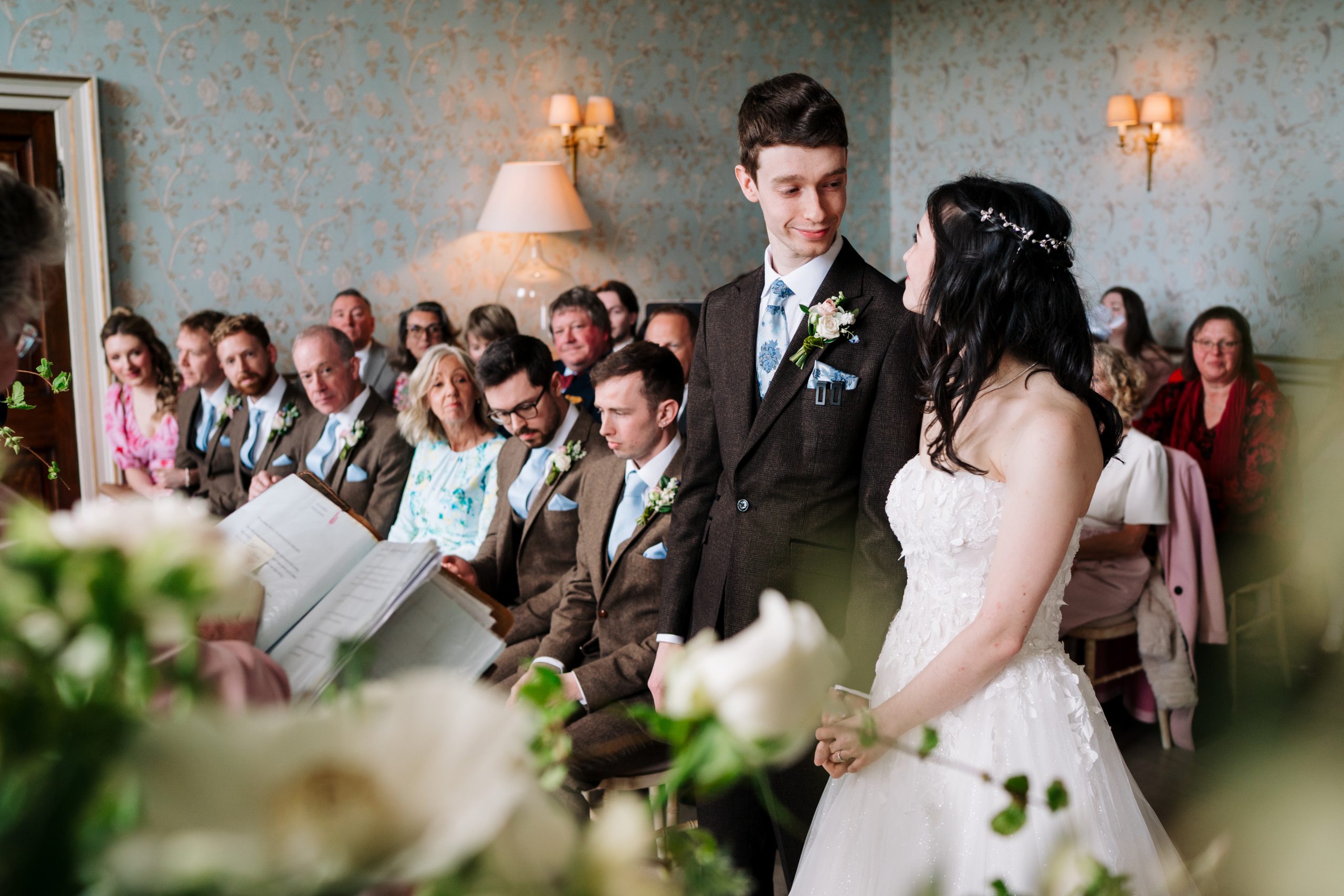 Bride in a white lace wedding dress and groom in a brown suit stand before seated guests in front of blue wallpaper, with warm lamps glowing.